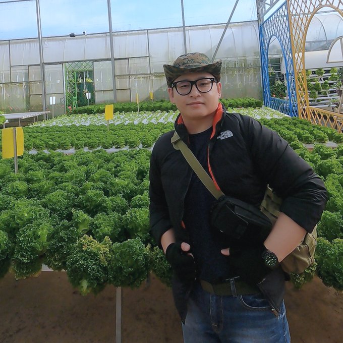 A man wearing a camouflage cap, glasses, and a black North Face jacket stands inside a greenhouse with rows of green lettuce growing in hydroponic beds behind him.