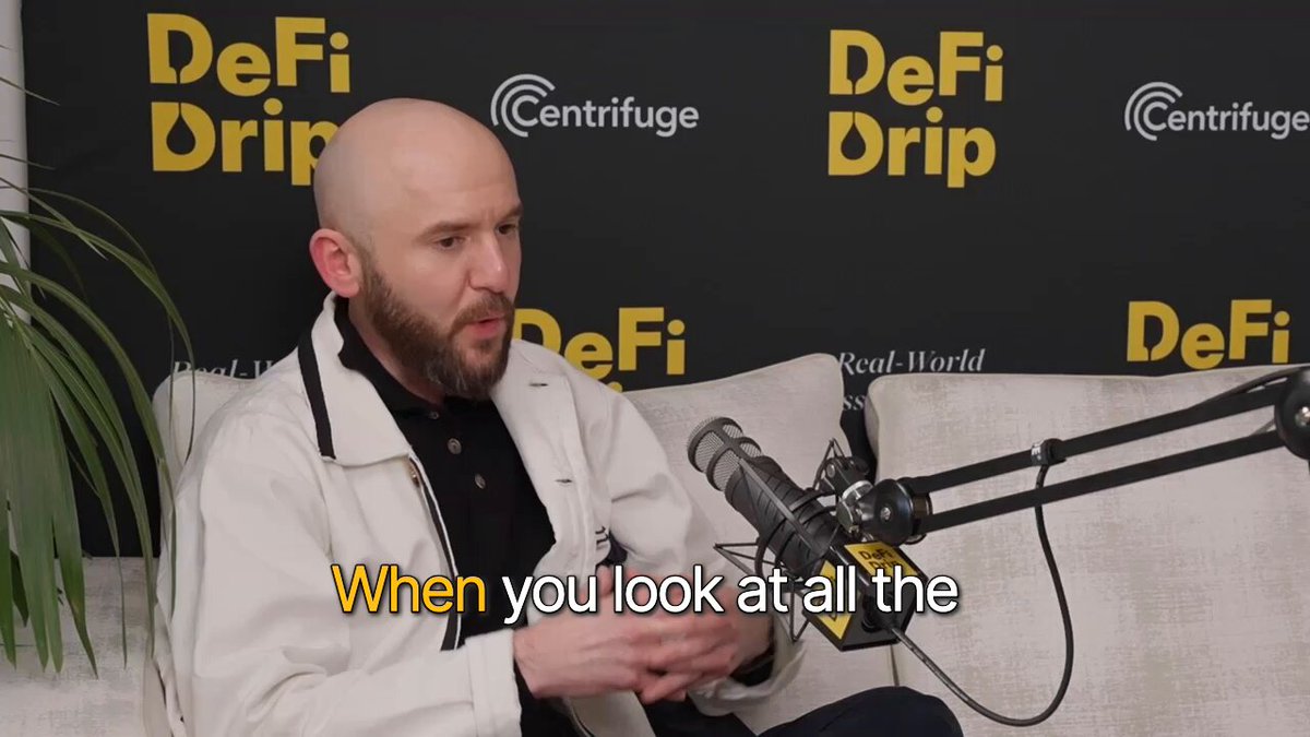 The image shows a bald man wearing a white coat sitting on a couch with his arms crossed, looking at something off-camera while holding a microphone. The background features a black and yellow sign that reads "Defi Dip" and "Centrifuge".