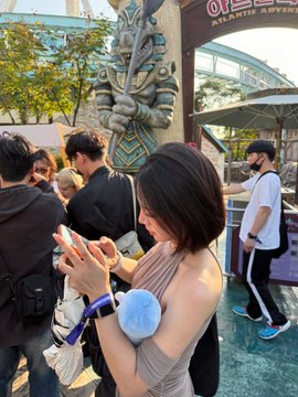 A woman in a tan top stands in the crowd at Universal Studios Japan, checking her phone while wearing a lanyard, with the entrance sign partially visible in the background. The scene reflects the crypto worker's post about juggling work messages and meetings during leisure activities and side events.