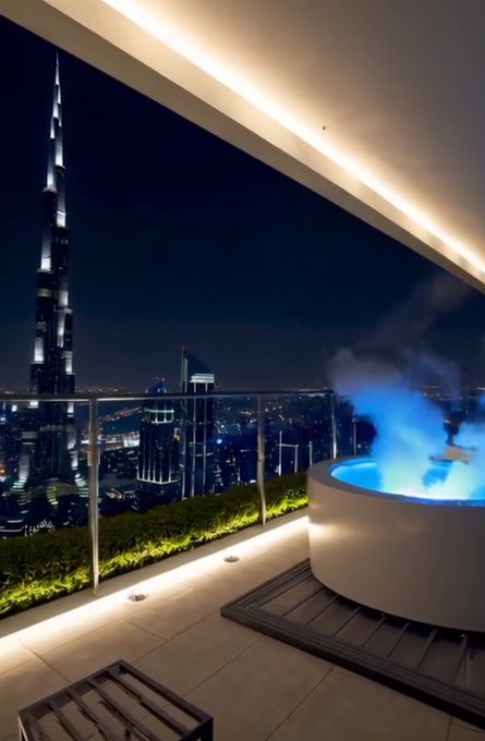 A luxurious rooftop terrace at night features a glowing blue hot tub emitting steam, set against the stunning backdrop of the Burj Khalifa and Dubai skyline. The image evokes a "lock in together" mindset of high-level success, visually representing the aspirational lifestyle the poster is channeling in their call for fellow crypto traders to "GM" (good morning) and stay focused.