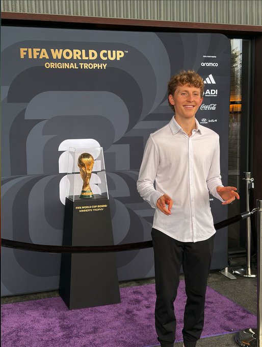 A young man with curly blonde hair, wearing a white button-up shirt and black pants, stands smiling beside the FIFA World Cup Original Trophy on display, with the backdrop reading "FIFA WORLD CUP ORIGINAL TROPHY" alongside sponsor logos including Aramco, Adidas, Coca-Cola, and Kia. The image captures a moment from his New York World Cup Final experience hosted by the $TRUMP Coin Club, showcasing one of the exclusive private events from the unforgettable weekend.