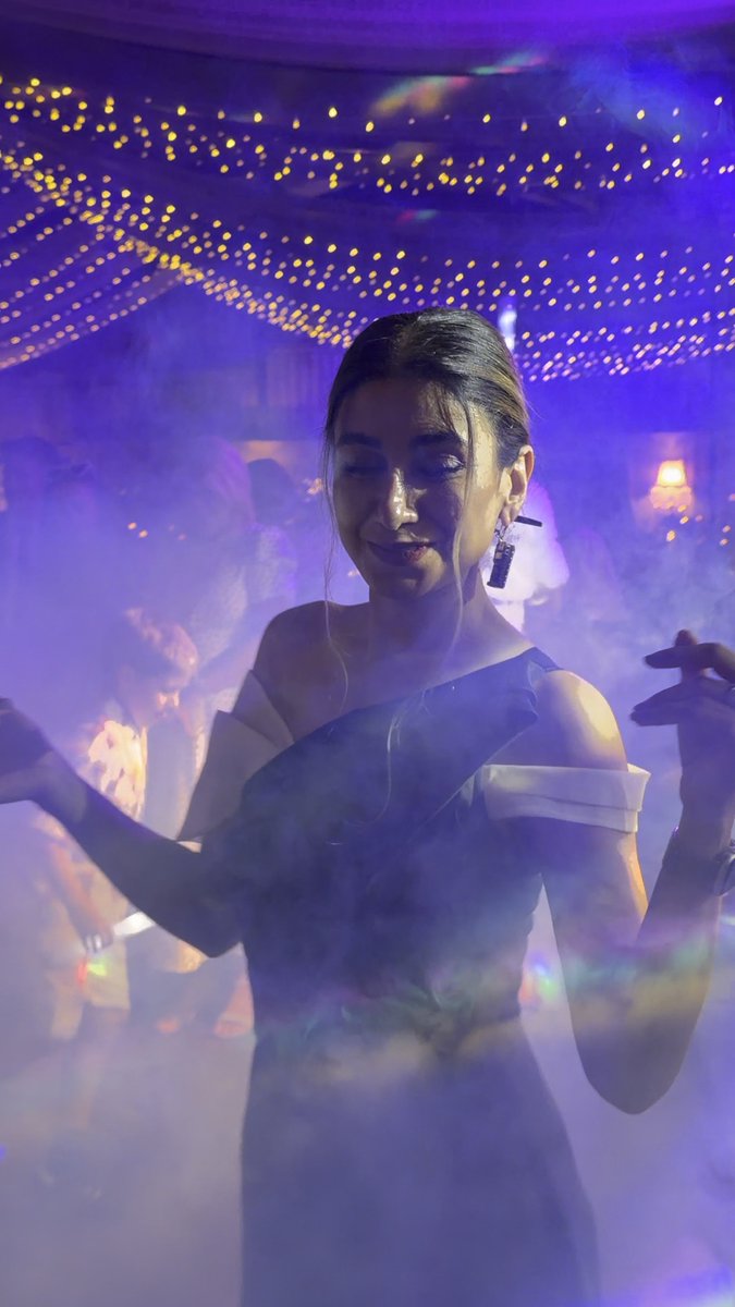 A woman with her hair up dances or poses in a smoky, purple-lit venue with string lights on the ceiling, wearing an off-shoulder dress and statement earrings.
