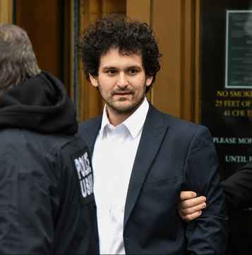 A man with curly dark hair, identified by context as Sam Bankman-Fried (SBF), is escorted by what appears to be law enforcement, wearing a dark suit jacket and white shirt with a "PRESS" badge visible on an adjacent jacket. The image relates to the post's discussion of SBF-linked donations to UK Defence Secretary Wes Streeting, as SBF is the disgraced FTX founder whose financial connections are at the center of the donation controversy.