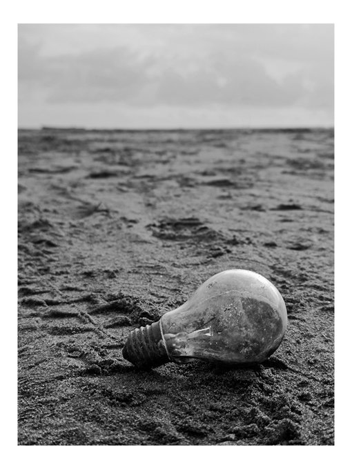 Black and white photo of a broken lightbulb lying on textured sandy ground.