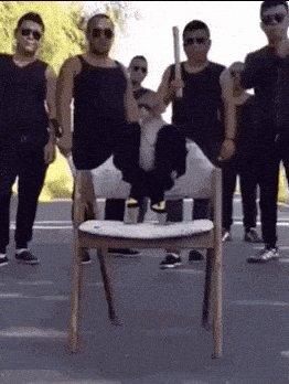 The image shows a man sitting on an empty chair, holding a baseball bat over his head as if he's about to hit something or someone. The scene takes place outdoors with several other people standing around him, some of whom are wearing sunglasses. There is also a bench in the background.