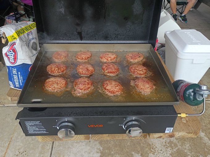 Twelve round sausage patties are sizzling on a black flat-top VEVOR griddle outdoors, with a bag of Kingsford charcoal and a white cooler nearby.