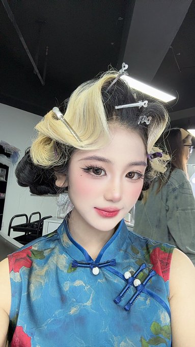 A young woman with dramatic makeup poses for a selfie while her hair is being styled in curlers and clips at a salon, wearing a blue floral cheongsam-style top with traditional knot buttons.