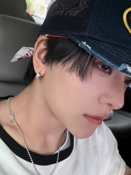 A close-up selfie of a young man wearing a dark baseball cap with a yellow logo, a white t-shirt with black trim, a silver chain necklace, and diamond earrings.