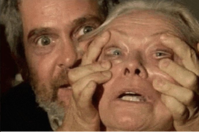 A man with wide, startled eyes holds his hands up to a woman's face, spreading her eyes open in an exaggerated expression of shock or panic. The image humorously conveys the post's caption "look at this, bitcoin," as a visual metaphor for the stunned reaction people might have when seeing Bitcoin's price or news.