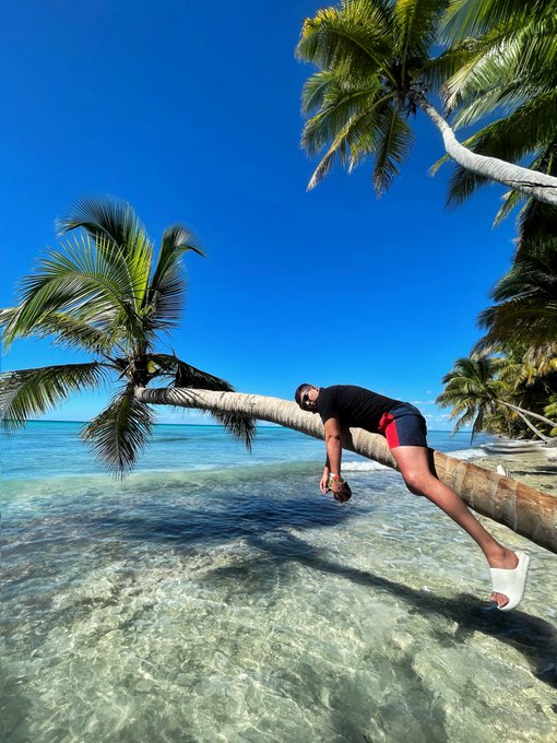 A man wearing a black t-shirt and red shorts hanging horizontally off the trunk of a palm tree leaning over clear turquoise ocean water under a bright blue sky.