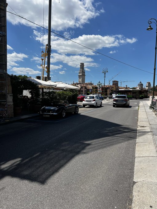A sunny street scene featuring a vintage black convertible and other cars parked near a plaza, with a historic tower visible in the distance under a cloudy blue sky.