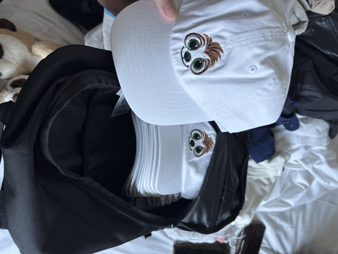 A hand holding a white baseball cap with an embroidered wide-eyed cartoon face design, with several identical caps stacked inside an open black bag.