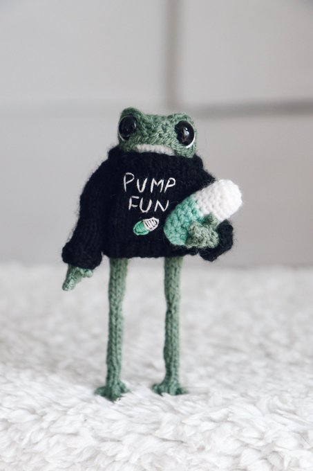A crocheted frog wearing a black sweater with "PUMP FUN" written on it stands on a white blanket, holding a small crocheted item, responding to the post's request to knit a frog for @Pumpfun.