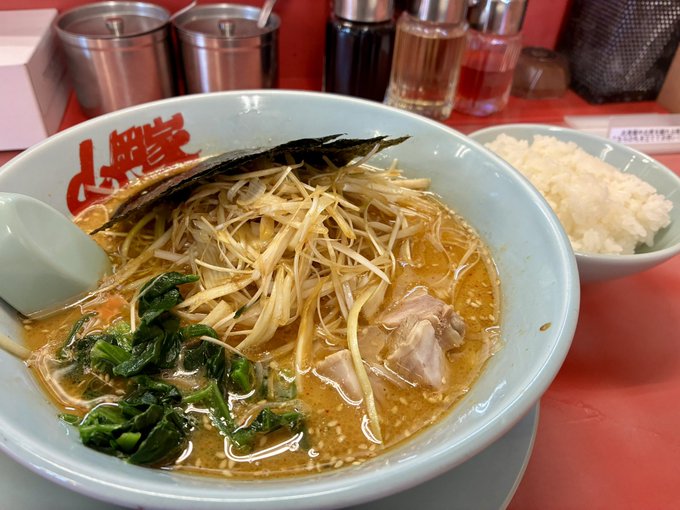 A bowl of ramen with noodles, bean sprouts, greens, and meat in broth, served with a side of white rice at a restaurant with red table and condiment bottles in the background.