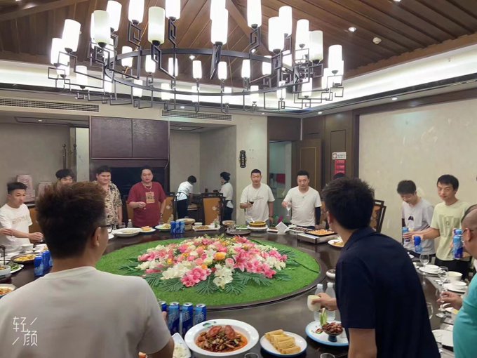 A group of men stand around a large, round dining table featuring a floral centerpiece and various dishes in a restaurant.