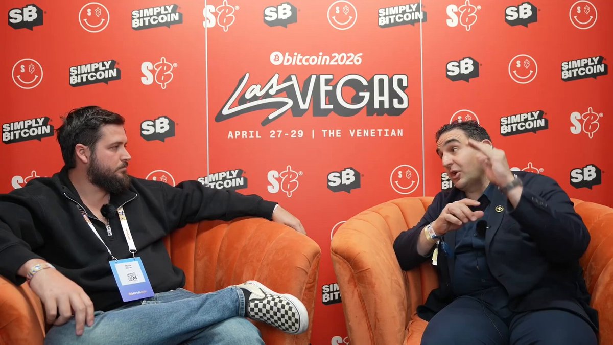 The image features two men sitting in chairs facing each other, with a large red backdrop behind them. One man has a name tag on his shirt that reads "Aydin Kilic", while the other man wears a suit and tie. The background includes text related to Bitcoin and cryptocurrency, such as "Bitcoin" and "LAS VEGAS". The image also contains a caption about Bitcoin's recent convertible note offering and how it is being deployed for mining and AI computing.