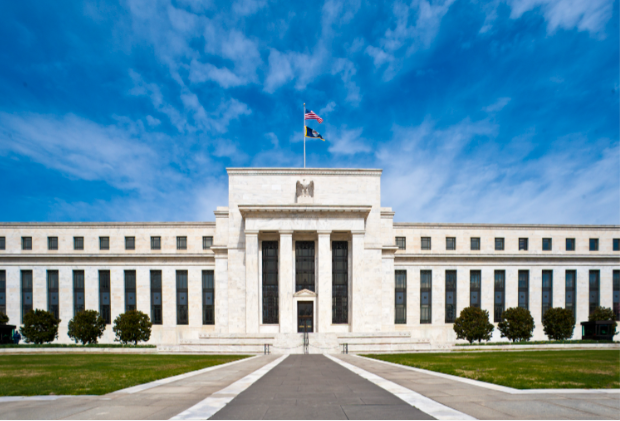 A photograph of the Federal Reserve building with its iconic eagle emblem above the entrance and the American flag flying on top, set against a bright blue sky with scattered clouds.
