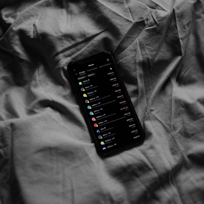 A smartphone resting on gray bedding displays an OKX "All Crypto" Markets list with tokenized U.S. stocks including NVDA ($285.12), INTC ($984.44), TSM, MRVL, and others available for 24/7 trading. The image visually reinforces the post's message by showing the mobile Wallet interface where users can trade AI-related tokenized equities anytime, without a traditional brokerage account.