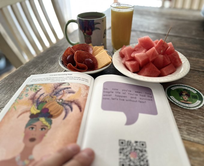 A person reads a colorful children's book at a wooden table, with a plate of cubed watermelon, a plate of pepperoni and cheese cubes, a mug, and a glass of orange juice nearby.