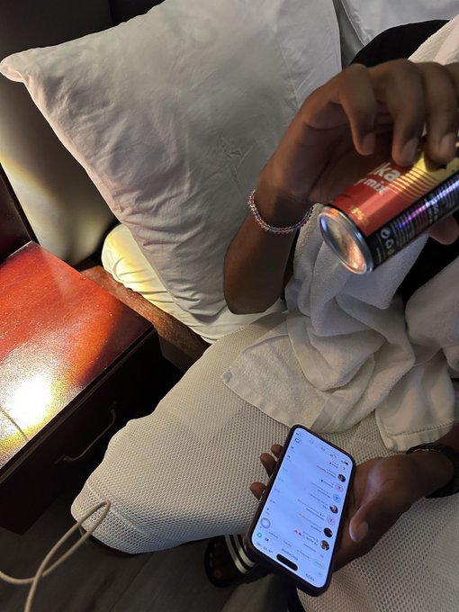 A first-person view shows hands holding a smartphone scrolling through social media while the other hand holds a gold and black beverage can, with pillows and a nightstand visible in the background.