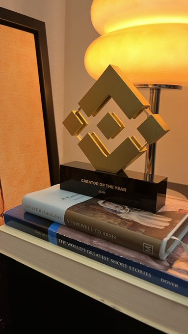 A golden "Creator of the Year" Binance Award trophy rests atop a stack of books—including Hemingway's *A Farewell to Arms* and *The World's Greatest Short Stories*—with a warm lamp glowing in the background, symbolizing the creator's gratitude to Binance for the recognition.