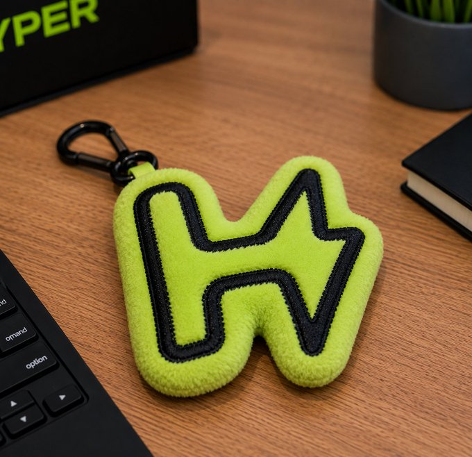 A neon green fuzzy keychain shaped like the letter "H" with black outline stitching sits on a wooden desk next to a keyboard and notebook, near a "HYPER" branded box.