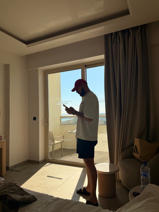 A man wearing a red cap and dark shorts stands on a balcony looking at his phone, with a stunning ocean view in the background. The "Gm friends 🖤" caption captures a relaxed morning vibe as he enjoys a peaceful start to the day in what appears to be a hotel or resort room.
