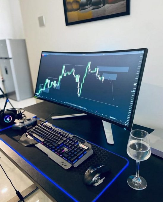 A curved monitor displays a bullish green candlestick chart with a drawn upward arrow, paired with a sleek gaming keyboard, mouse, and a glass of water on a desk. The upward-trending chart visually embodies the post's hopeful message that "Joy is coming, Money is coming," symbolizing financial growth and optimism.