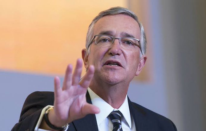 A photograph of an older man with gray hair and rimless glasses, wearing a dark suit and striped tie, gesturing with his raised right hand while speaking.