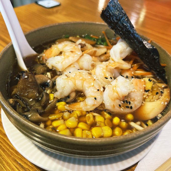 The image features a bowl of shrimp soup with noodles, vegetables, and corn. The bowl has a white plastic spoon inside it. There are also some mushrooms mixed into the dish. The photo appears to be taken at a restaurant or dining establishment where people can enjoy their meal in a comfortable setting.