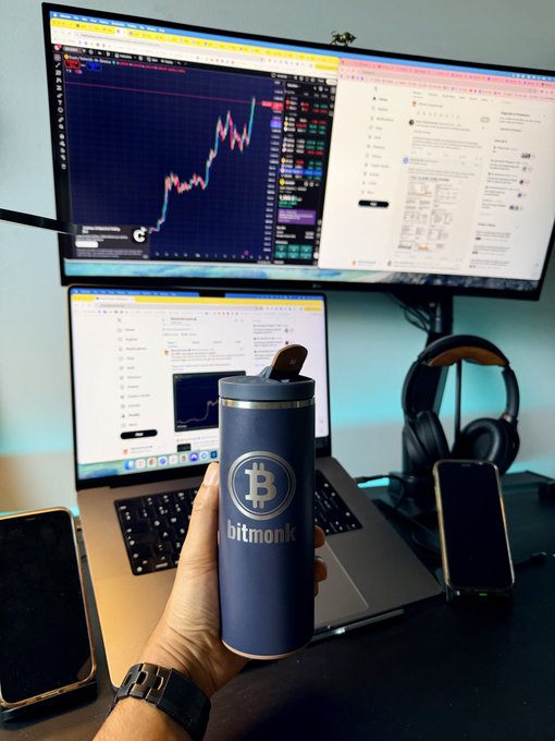 A hand holds a navy "bitmonk" Bitcoin-branded tumbler in front of a dual-screen setup showing a BTC/USD chart on TradingView and crypto market news, complementing a morning market update noting BTC around $76K and ETH near $2.4K post-Fed recovery.