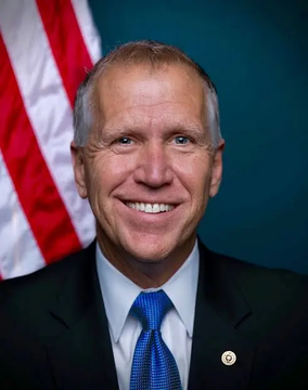 An official portrait of Sen. Thom Tillis, a smiling man in a dark suit with a blue tie and a Senate lapel pin, posed in front of an American flag. The image accompanies a breaking news post where Tillis urges a Clarity Act vote next week, emphasizing the need to establish an ethics baseline beforehand.