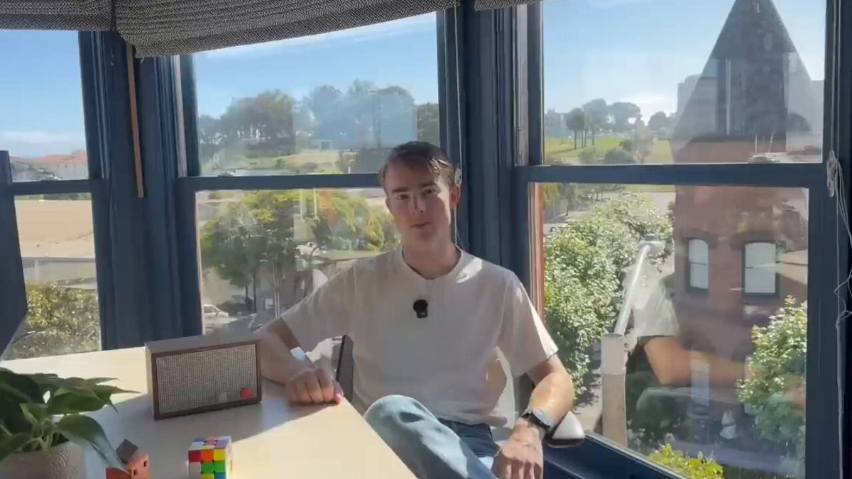 The image shows a man sitting at a desk with a window behind him, looking out onto an outdoor view. The man has a Rubik's cube next to him on the table. There are also several other objects present in the scene, including a potted plant and a radio. The post seems to be about someone giving their own radio station to AI models for six months, with each model losing its mind in different ways during that time.