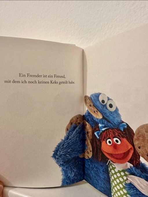 A Cookie Monster plush and a red-haired Sesame Street Muppet pose beside an open book featuring the German text: *"Ein Fremder ist ein Freund, mit dem ich noch keinen Keks geteilt habe."* ("A stranger is a friend with whom I haven't yet shared a cookie.") The cookie-themed quote ties into the "GM fam" post, highlighting a friendly, welcoming morning vibe.