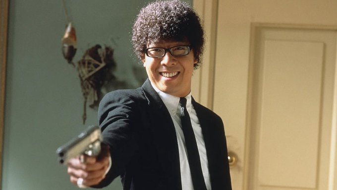 A man with a curly afro wig and black glasses, wearing a black suit and tie, smiles while pointing a small silver pistol at the camera in an indoor setting.