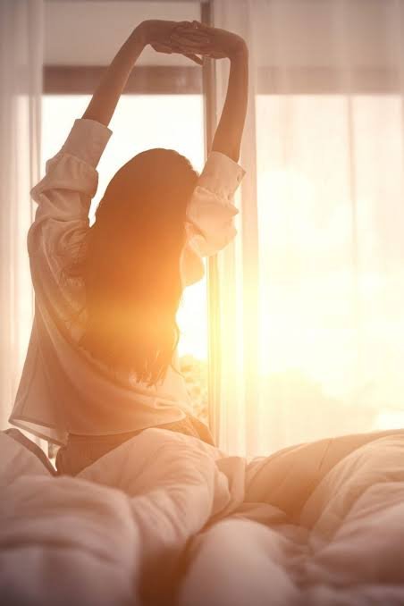 A silhouetted woman stretches her arms above her head while sitting on a bed, bathed in warm, golden sunlight streaming through sheer curtains.