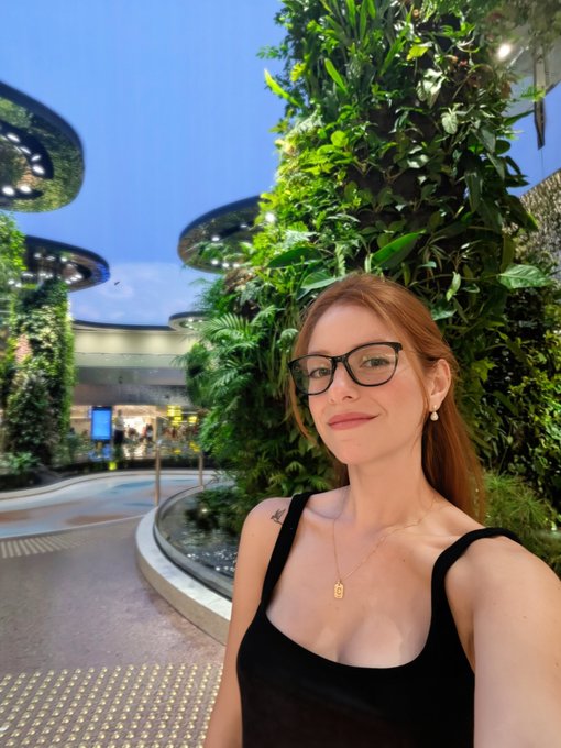 A woman with red hair and black glasses takes a selfie in what appears to be a modern building with a lush green living wall and curved architecture in the background.