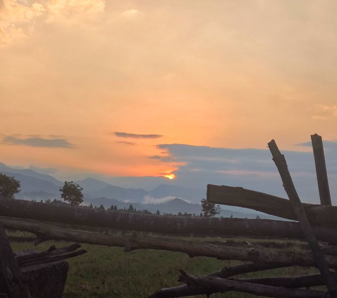 A scenic landscape at sunset with the sun dipping behind distant mountains, viewed past a rustic wooden fence in the foreground.