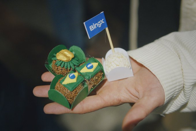 A hand holds small paper-crafted treats themed with Brazilian flags, a golden cocoa pod, and a Bingx-branded flag on a toothpick.