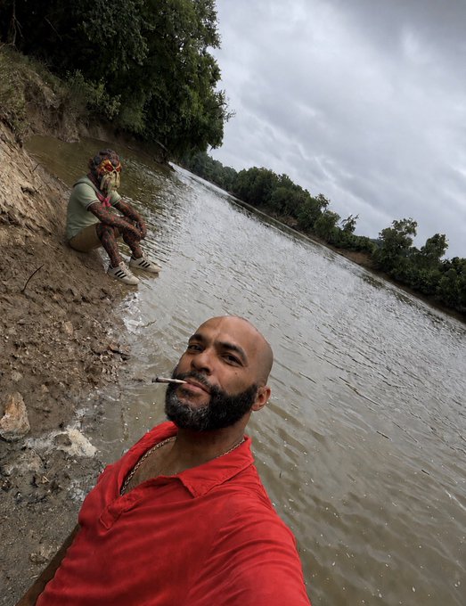 The image shows a man wearing a red shirt sitting by a river, holding a cigarette and looking at his phone. The photo has been edited with a filter that gives it a vintage appearance. The caption of the image reads "GMGMGM Some days you just gotta unplug and wait for ApeFest……." This suggests that the man is taking a break from his daily routine to enjoy some leisure time by the river, possibly capturing this moment on camera or sharing it with friends online.
