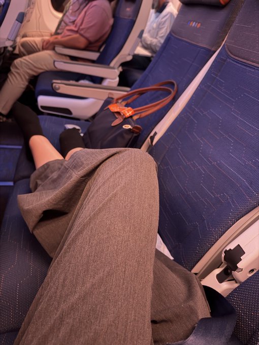 A passenger sits in a blue airplane seat with crossed legs and a brown handbag on the adjacent seat, while other passengers are visible in the background.