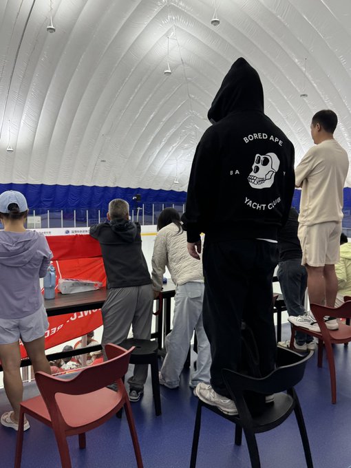 A group of people stand on chairs at an indoor sports arena, with one prominently wearing a black "BORED APE YACHT CLUB" hoodie, embodying the pride and community spirit of being part of the iconic NFT club. The elevated seating captures the moment as fans gather to celebrate their shared membership in the legendary digital collective.
