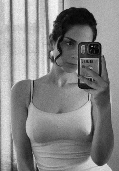 A woman takes a black-and-white mirror selfie wearing a fitted tank top, holding a smartphone displaying "I BELIEVE" on its case. The image reflects a confident, minimalist aesthetic that complements the casual "gm" (good morning) greeting, suggesting a calm and introspective start to the day.