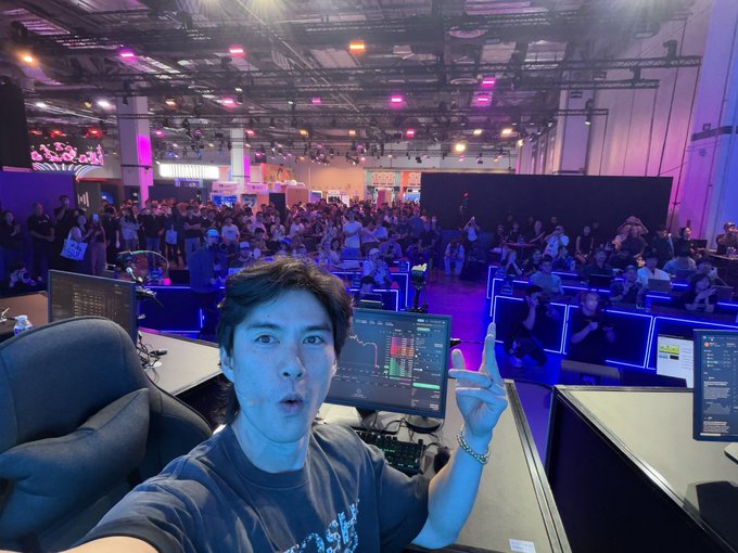 A man gestures excitedly at a trading floor with rows of seated spectators watching multiple monitors displaying charts and order books, capturing the quiet moment of an overnight US session before Asian markets open.