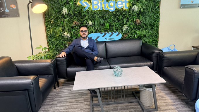 A man in a suit sits on a black leather couch in a Bitget office, with the Bitget logo visible on a plant-covered wall behind him.