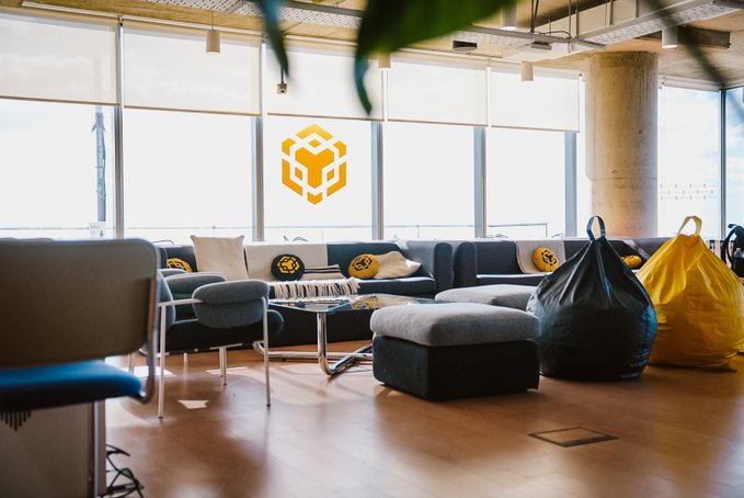 A modern, sunlit lounge area featuring floor-to-ceiling windows adorned with a large gold BNB (Build N Build) cube logo, gray sectional sofas with yellow BNB-themed pillows, a coffee table, and black and yellow bean bags set up for a casual gathering.

The image illustrates the inviting community event spaces referenced in the post, showcasing the kind of comfortable BNB Guilds meetup venue where local crypto enthusiasts could gather for networking and discussions.