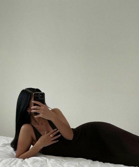 A woman with long dark hair in a black dress lies on a white bed, taking a mirror selfie with her phone that obscures her face.