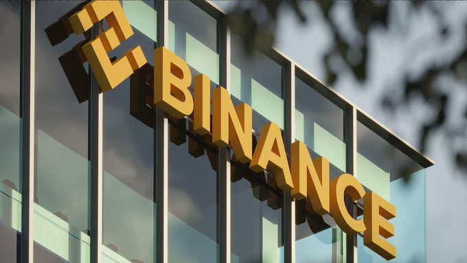 A photograph of a modern glass building facade displaying large, 3D yellow letters spelling out 'BINANCE' alongside its diamond-shaped logo, with green tree leaves visible in the upper right corner.