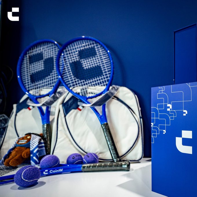Two blue tennis rackets lean against white bags alongside purple tennis balls and a small mascot plush, with one racket branded "CoinW" and a matching blue box displaying the company's "C" logo nearby. The image visually merges sportsmanship with the trading platform's branding, reinforcing the "Serve. Trade. Repeat." tagline by pairing athletic precision with CoinW's financial presence.