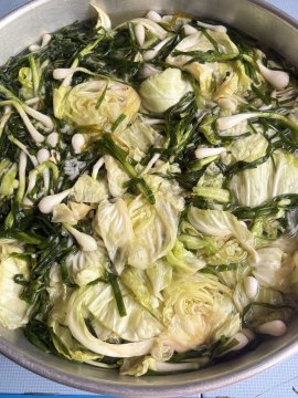 A close-up photograph of a metal bowl filled with a soup or stew containing cabbage, leafy greens, and enoki mushrooms.